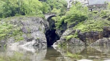 Traffiume, Cannobio, Piemont, İtalya 'da yemyeşil yemyeşil ile çevrili Orrido di Sant' Anna 'daki sakin bir nehrin üzerindeki antik taş köprünün yanında çan kulesi olan tarihi bir kilisenin resmi.