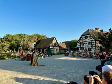 Ungersheim, France - August 16 2024 : Traditional village festival performance at Ecomusee d'Alsace with a stilt walker and golden-robed performer entertaining a diverse summer crowd in open-air park clipart