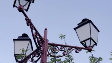 Alacakaranlıkta açık mavi gökyüzüne karşı yeşil yapraklarla çevrili üç fenerli eski tip demir sokak lambası klasik tasarım ve doğanın karışımını sergiliyor.