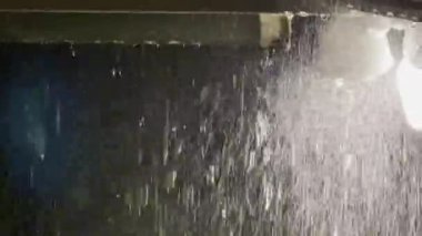 Gece vakti açık havada parlak bir ışık altında yağan yağmur damlaları ve su akıntıları çarpıcı bir yağmur sahnesinde karanlık bir zemin üzerine vurgulanıyor.