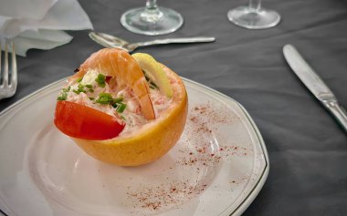 Elegant seafood shrimp cocktail served in halved grapefruit with creamy dressing, garnished with fresh chives, tomato, and lemon slices on a white plate with cutlery in a formal dining setting clipart