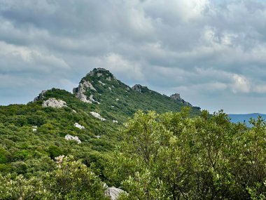 View of Col de la Pousterle and Montferrand Castle Among Lush Mediterranean Forest and Limestone Peaks Under a Dramatic Clouded Sky Near Pic Saint-Loup, Southern France clipart