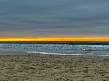 Golden Sunrise Over the Mediterranean Sea at Palavas-les-Flots, France, with a Small Sailboat on the Horizon, Gentle Waves, Sandy Beach, and Dramatic Overcast Clouds clipart