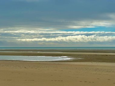 Baie d 'Authie' nin dalgaların çekildiği engin ve sakin bir manzarası, sığ gelgit havuzlarında yüzen yalnız beyaz bir kuş, kıyı şeridi boyunca uzak insan siluetleri, ve dramatik bulutlarla dolu bir gökyüzü.