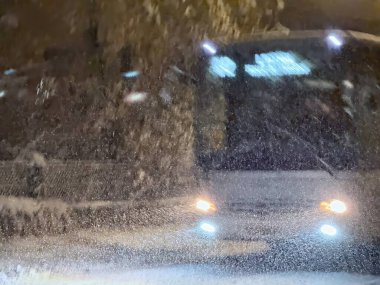 Gece ağır kar yağışı altında giden bir otobüsün bulanık görüntüsü, farları açık ve parlak ekran kar kaplı ön camdan zar zor görülebiliyor, atmosferik bir kış manzarası yaratıyor.