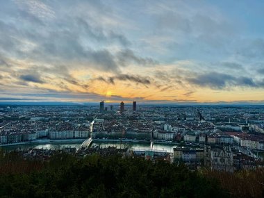 Modern gökdelenlerin ve tarihi çatıların siluetleri ile Lyon şehri üzerinde geniş panoramik gündoğumu manzarası, dramatik bulutlarla dolu bir gökyüzünün altında Fourviere tepesinden görülüyor.