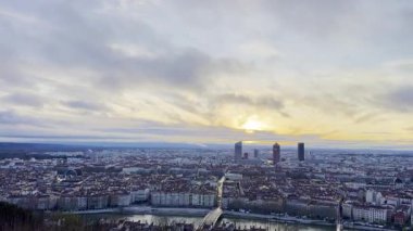 Modern gökdelenlerin ve tarihi çatıların siluetleri ile Lyon şehri üzerinde geniş panoramik gündoğumu manzarası, dramatik bulutlarla dolu bir gökyüzünün altında Fourviere tepesinden görülüyor.