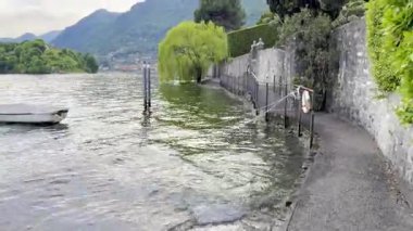 Lenno, İtalya - 2 Mayıs 2025: Lenno ve Ossuccio 'daki huzurlu göl gezintisi, düzgün budanmış ağaçlar, sahil bankları, dağ manzaraları, renkli köyler ve Como Gölü üzerindeki manzaralı perspektifler