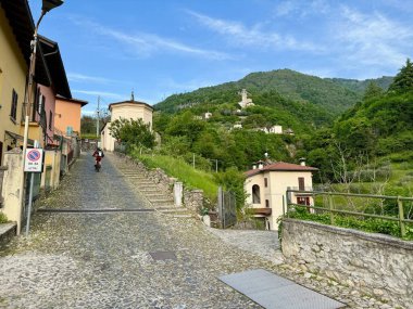 Lenno, İtalya - 3 Mayıs 2025: Lenno Ossuccio 'da dik kaldırım taşı caddesi, bisikletçi renkli evlerin, tarihi şapelin ve yeşil yamaçların önünden geçerek parlak bahar gökyüzünün altında