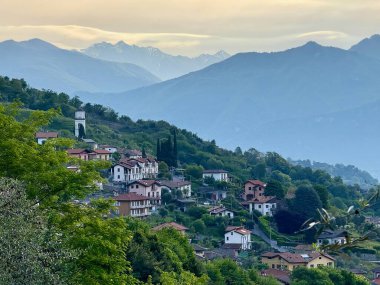 Sabahın erken saatlerinde Como Gölü ve etrafındaki dağların Sacro Monte di Ossuccio, İtalya 'daki hac yolundan zeytin ağaçları, çatılar ve bulutları delen yumuşak güneş ışınlarıyla manzarası.