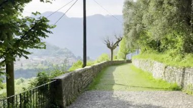 Sabahın erken saatlerinde Como Gölü ve etrafındaki dağların Sacro Monte di Ossuccio, İtalya 'daki hac yolundan zeytin ağaçları, çatılar ve bulutları delen yumuşak güneş ışınlarıyla manzarası.