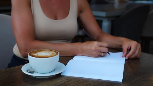 一个无法辨认的女人坐在咖啡店的桌子旁 一边喝咖啡 一边写着写信息的笔记本 高质量的4K镜头 — 图库视频影像