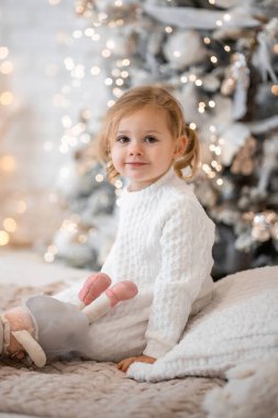 Beyaz elbiseli, çok hoş bir sarışın kız çocuk yatağında oturuyor ve evin parlak iç kısımlarındaki Noel ağaçlarının arka planında gülüyor. Yüksek kalite fotoğraf