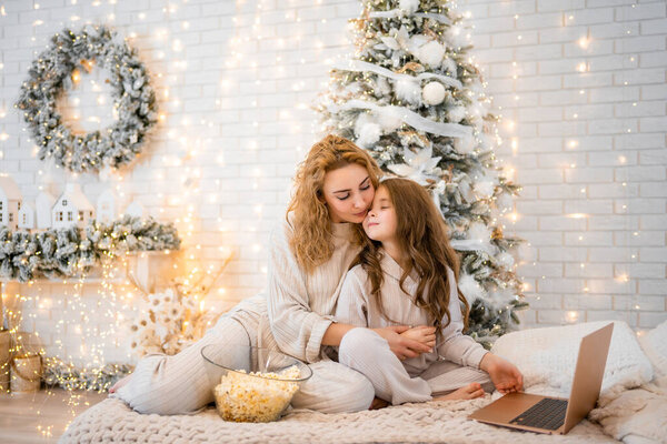 Young woman with child baby girl 7 years old in sleepwear watching film and eating popcorn on the bed in Christmas decorated home. High quality photo