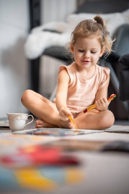 Little girl sits on the floor at home and draws with paints and brushes in a coloring book. Early childhood creativity and education. High quality photo
