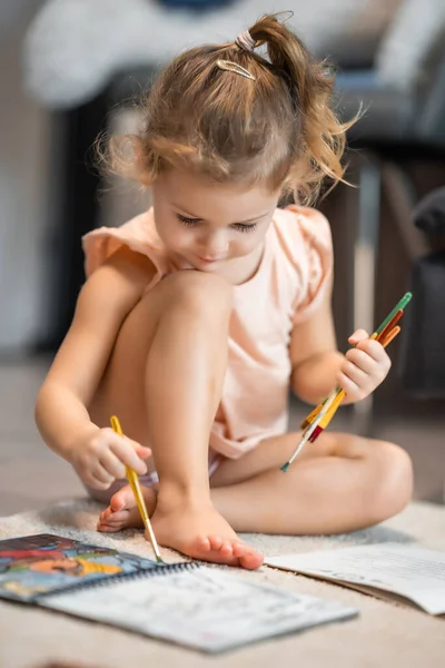 Little girl sits on the floor at home and draws with paints and brushes in a coloring book. Early childhood creativity and education. High quality photo