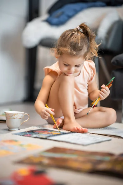 Little girl sits on the floor at home and draws with paints and brushes in a coloring book. Early childhood creativity and education. High quality photo