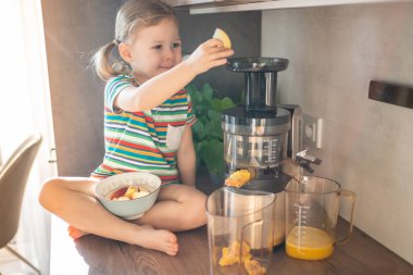 Little girl making fresh juice sitting on the table in home kitchen. High quality photo