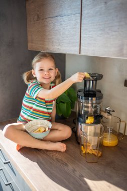 Little girl making fresh juice sitting on the table in home kitchen. High quality photo