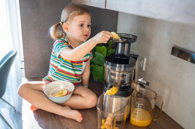 Little girl making fresh juice sitting on the table in home kitchen. High quality photo