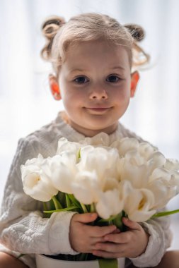 Lale çiçeği buketiyle pencerenin kenarında oturan küçük kız. Mutlu çocuk, içeride. Anneler günü, sevgililer günü ya da doğum günü konsepti. Yüksek kalite fotoğraf