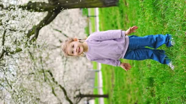 小さな女の子は咲くリンゴの木の下に立っている 風が吹き 花の花びらがヨーロッパのプラハ公園の雪のように飛びます 高品質4K映像 — ストック動画