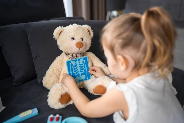 Little girl playing doctor with toys and teddy bear on the sofa in living room at home. High quality photo