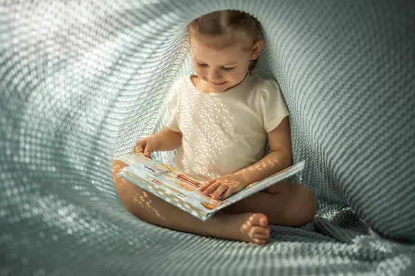 Little girl enjoying of review a book under blue knitted plaid in sunny morning. High quality photo