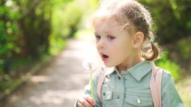 Mutlu bir kızın portresi. Çocuk bahar günü gülümsüyor. Çocuk baharın tadını çıkarıyor. Güneşli bir gün. Sarışın kız karahindiba üflüyor. Açık hava, kapatın. Yüksek kalite 4k görüntü