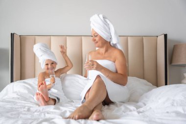 Mutlu aile annesi ve küçük kızı sabahlıklar ve havlular içinde evde spa günü boyunca bardakla su içip gülüyorlar. Yüksek kalite fotoğraf
