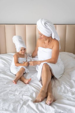 Mutlu aile annesi ve küçük kızı sabahlıklar ve havlular içinde evde spa günü boyunca bardakla su içip gülüyorlar. Yüksek kalite fotoğraf