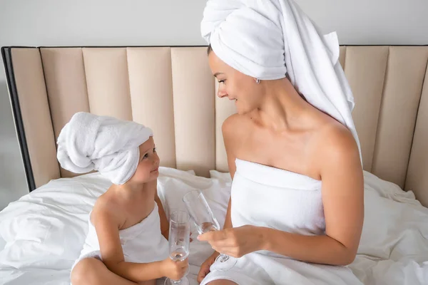 Happy family mother and little daughter in dressing gowns and towels laugh and drinking water with glasses during spa day at home. High quality photo