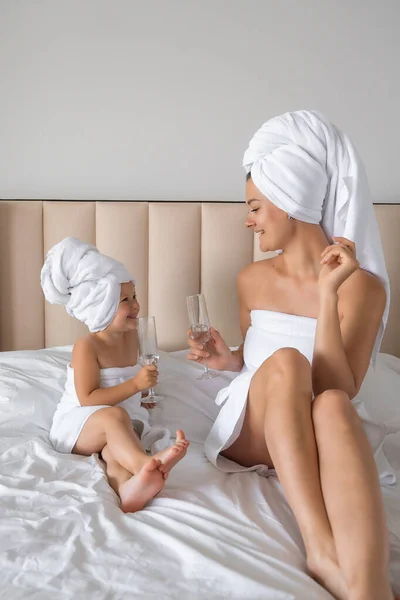 Happy family mother and little daughter in dressing gowns and towels laugh and drinking water with glasses during spa day at home. High quality photo