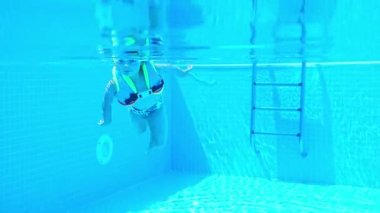 Gözlüklü ve havuzda yelek giymiş su altında yüzen küçük bir kızın portresi. Yaz tatili. Yüksek kalite 4k görüntü