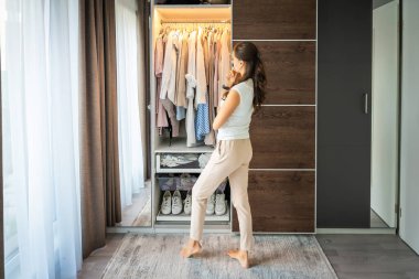 Uzun kıvırcık kıvırcık saçlı, günlük kıyafetlerle gülümseyen genç bir kadın gardıropta duruyor ve çeşitli elbise ve ayakkabılarla rafların yakınındaki askılardan kıyafet seçiyor. Yüksek kalite fotoğraf