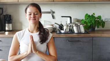 Genç bir kadın mutfakta oturmuş kirli bulaşıkların arka planı üzerine meditasyon yapıyor. Yavaşla, kendine dikkat et, yavaş yaşam konsepti. Yüksek kalite 4k görüntü