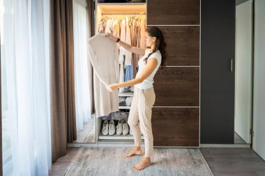 Uzun kıvırcık kıvırcık saçlı, günlük kıyafetlerle gülümseyen genç bir kadın gardıropta duruyor ve çeşitli elbise ve ayakkabılarla rafların yakınındaki askılardan kıyafet seçiyor. Yüksek kalite fotoğraf