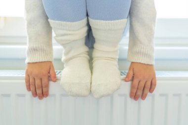 Sıcak beyaz çoraplı küçük bir kızın elleri ve ayakları. Çocuk pencerenin kenarında oturuyor ve ısıtma kaloriferinden ısınıyor. Evde, bir dairede ısıtma. Yüksek kalite fotoğraf