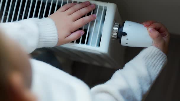 暖かい白いセーターの小さな女の子は窓に立って電池暖房のノブを回し 暖房ラジエーターから暖めます 自宅でアパートで暖房をしています 高品質の4K映像 — ストック動画