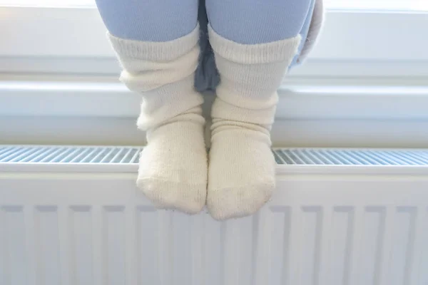 Feet of little girl in warm socks. Child sitting by window and warming up from the heating radiator. Heating in an apartment, at home. High quality photo
