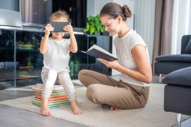 Küçük kız bir yığın çocuk kitabının üzerine oturur ve annesi kitap okurken akıllı telefondan çizgi film izler. Yüksek kalite fotoğraf