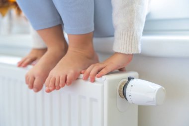 Küçük kızın elleri ve ayakları ısıtıcının üzerinde. Çocuk pencerenin kenarında oturuyor ve ısıtma kaloriferinden ısınıyor. Batarya ısıtma düğmesine odaklan. Evde, bir dairede ısıtma. Yüksek kalite fotoğraf