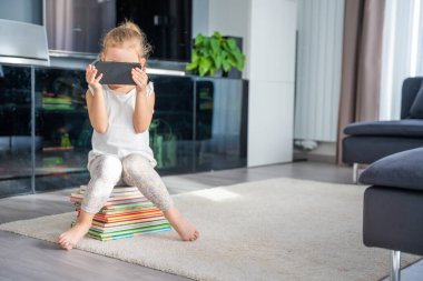 Şirin küçük kız bir yığın çocuk kitabının üzerine oturur ve akıllı telefondan çizgi film izler. Yüksek kalite fotoğraf