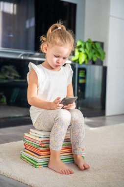 Küçük kız peri masalı kitaplarının üzerine oturmuş akıllı telefonuyla oynuyor. Yüksek kalite fotoğraf