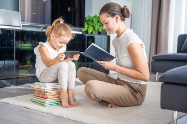 Küçük kız bir yığın çocuk kitabının üzerine oturur ve annesi kitap okurken akıllı telefonunu kullanır. Yüksek kalite fotoğraf