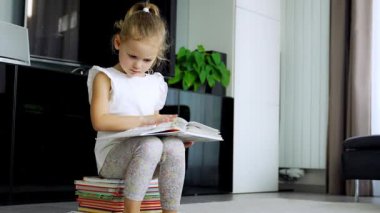 Küçük kız bir yığın çocuk kitabının üzerinde oturuyor ve dikkatle peri masallarıyla dolu bir kitabı karıştırıyor. Yüksek kalite 4k görüntü