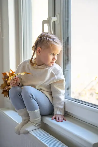 Little sad girl in warm sweater and socks sitting by window, holding autumn leaf and warming up from the heating radiator. Heating in an apartment, at home. High quality photo