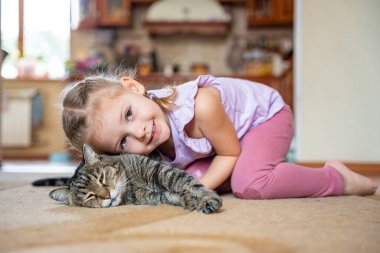 Gri kediyi kucaklayan ve oturma odasında halıya uzanan tatlı küçük bir kız. Yüksek kalite fotoğraf