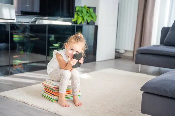 Gülümseyen küçük kız bir yığın çocuk kitabının üzerine oturur ve akıllı telefonunu kullanır. Yüksek kalite fotoğraf