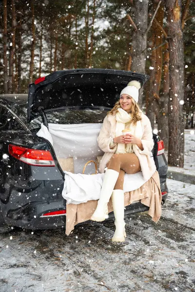 Young woman in woolen hat sits in trunk of the car. Girl have a fun in the car. Winter travel, snow-covered trees in the forest. Snowy weather. High quality photo